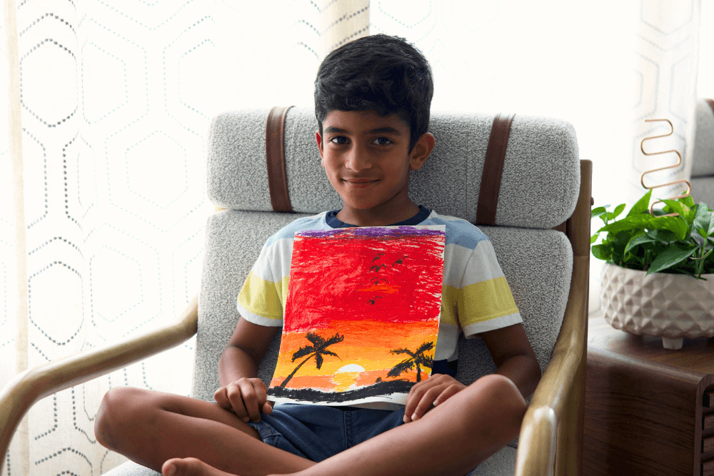 George proudly holding his vibrant sunset painting created with oil pastels. The artwork showcases bold shades of orange, pink, and purple blending beautifully across the sky, with silhouettes of trees in the foreground.