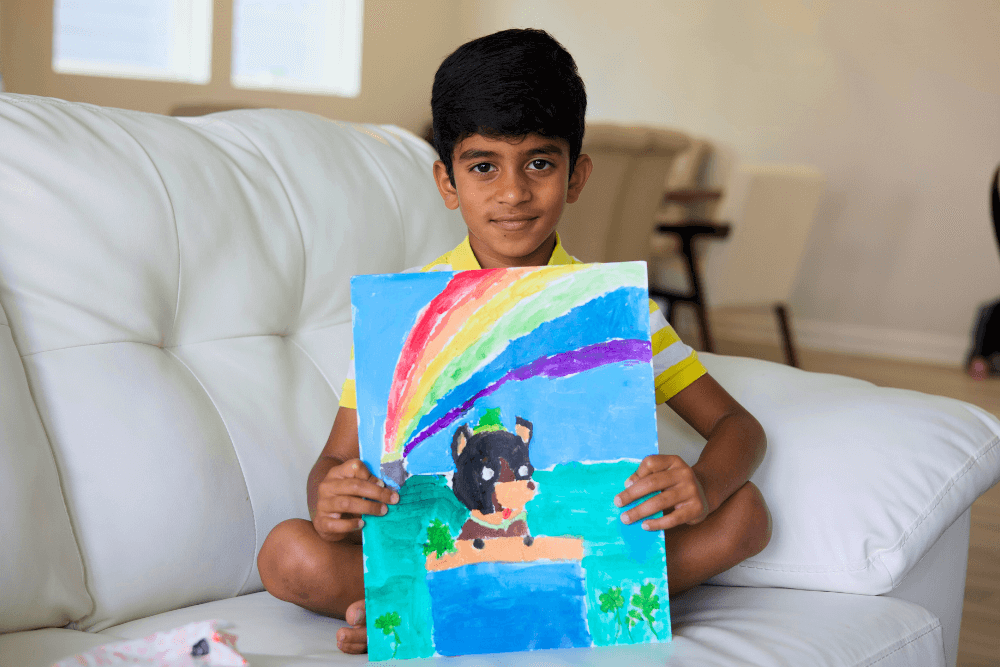 Young artist George proudly holds up his painting of a dog sitting in front of a vibrant rainbow.