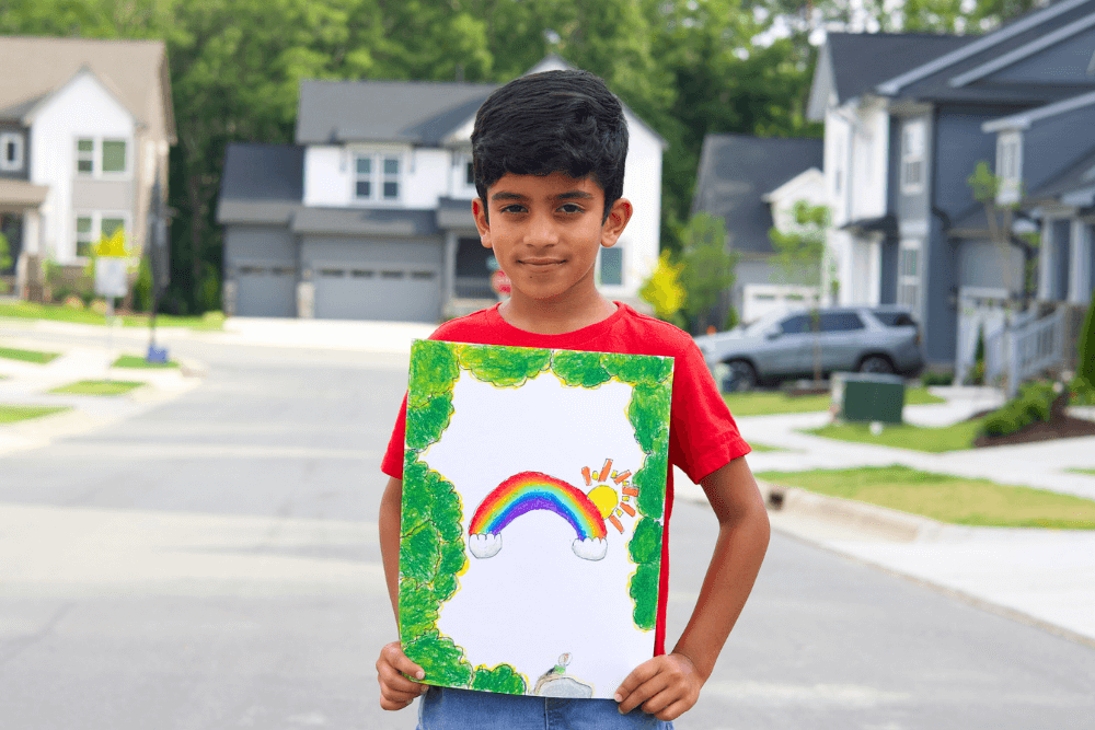 Young artist George proudly holds his vibrant oil pastel painting titled "FIND A RAINBOW," featuring a colorful rainbow arching across a serene landscape with a bright sky.