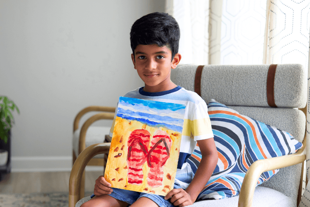 eorge smiling and holding his summer watercolor painting, featuring colorful slippers on a sandy seashore with gentle ocean waves in the background.