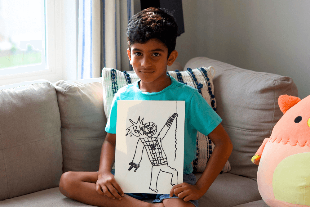 George proudly holding his hand-drawn picture of Spiderman, smiling with excitement. The drawing shows Spiderman in a dynamic pose, outlined with bold lines