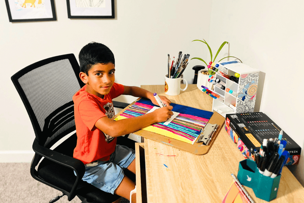 George sits at an art desk in his colorful art room surrounded by brushes, paints, and his vibrant artwork displayed on the walls.