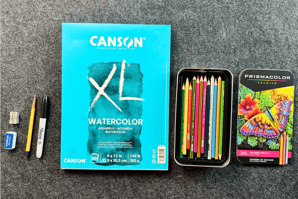 A set of Prismacolor colored pencils arranged, with a sketchbook and sharpener beside them.