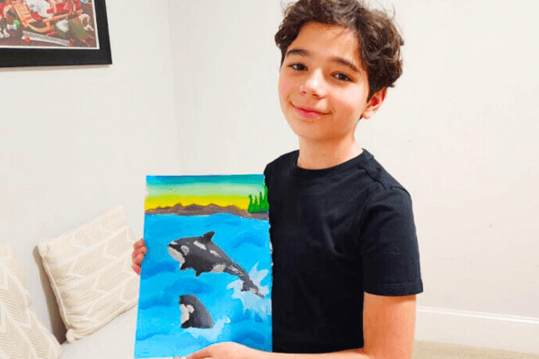 Kivanc holds up his vibrant watercolor painting of dolphins joyfully swimming in the ocean, created during the Summer Art Camp